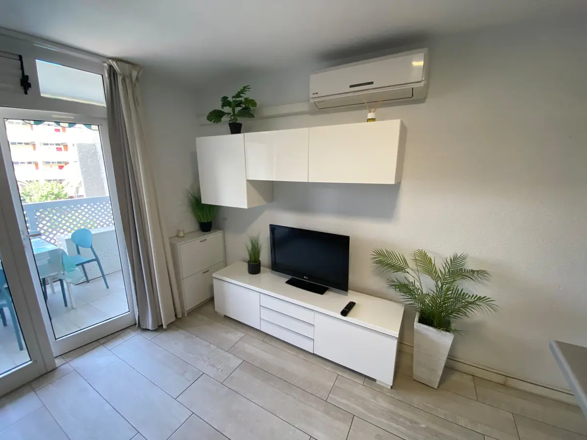 Helles Wohnzimmer mit weißem Möbel, Fernseher, Klimaanlage und Schiebetür zum Balkon